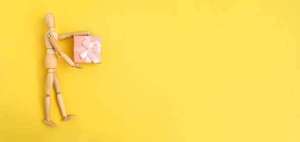Figura de madera sosteniendo una caja de regalo rosa. Fondo amarillo, espacio para copiar. Banner - Foto, Imagen