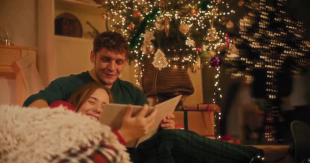 Ευτυχισμένο νεαρό ζευγάρι που βλέπει φωτογραφικό άλμπουμ στο σπίτι την παραμονή των Χριστουγέννων - Πλάνα, βίντεο