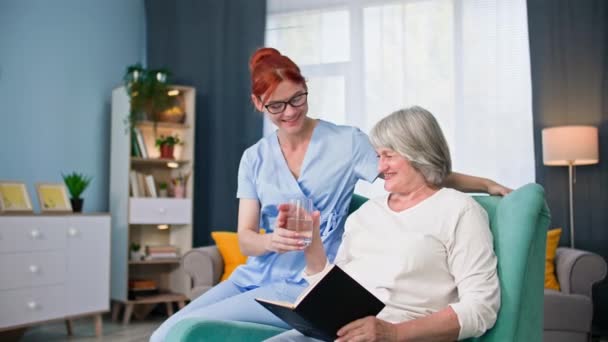 γηροκομείο, γυναίκα υπάλληλος με ιατρική στολή δίνει ένα ποτήρι νερό σε μια ηλικιωμένη γυναίκα στο άνετο δωμάτιο - Πλάνα, βίντεο