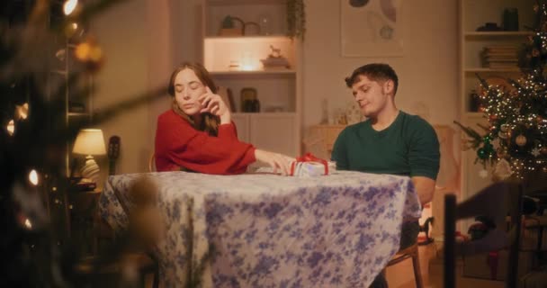 Mladý muž dává vánoční dárek pro přítelkyni, zatímco sedí v obývacím pokoji doma během dovolené - Záběry, video