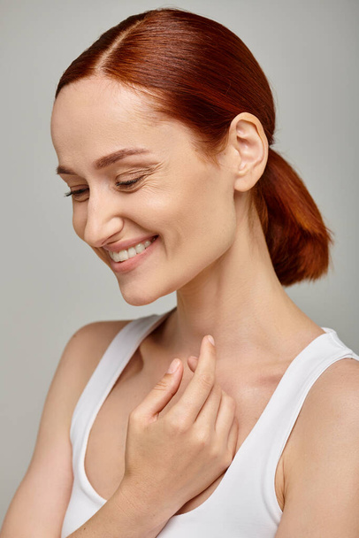 cheerful redhead woman in white tank top exuding joyful and healthy smile on grey background - Foto, Bild