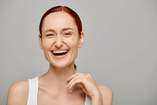 boldog és vörös hajú nő fehér tartály tetején egészséges mosollyal szürke háttér, nevetés - Fotó, kép