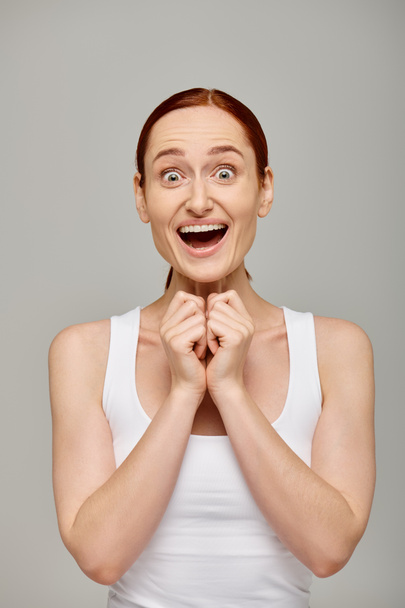 amazed and redhead woman in white tank top with healthy smile on a grey background, laughter - Photo, Image
