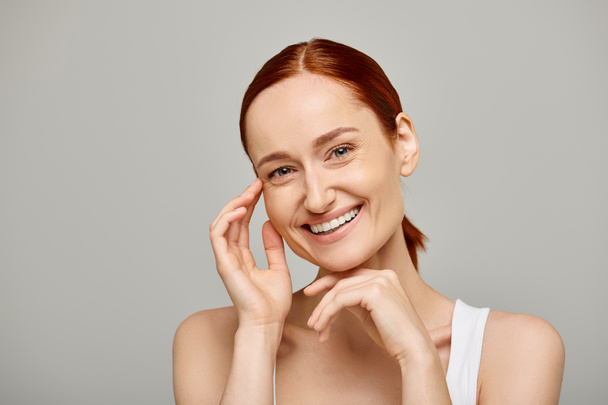 happy and redhead woman in white tank top looking at camera on grey background, healthy smile - Photo, Image
