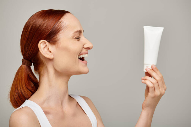 roodharige jonge en gelukkige vrouw in de 30 kijken naar buis met body lotion op grijze achtergrond, huidverzorging - Foto, afbeelding