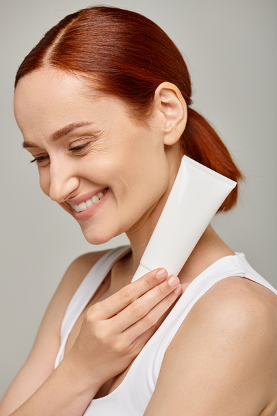cheerful young woman with red hair holding tube with body lotion on grey background, skincare - Photo, Image
