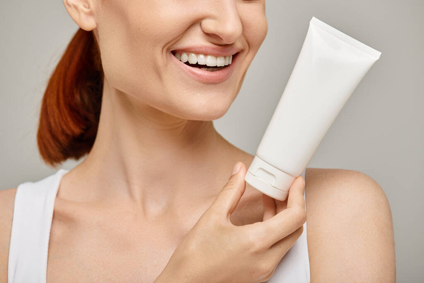 cropped view of cheerful woman with red hair holding tube with body lotion on grey background - Photo, Image