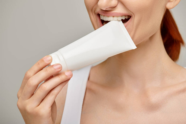 cropped view of cheerful woman with red hair biting tube with body lotion on grey background - Photo, Image