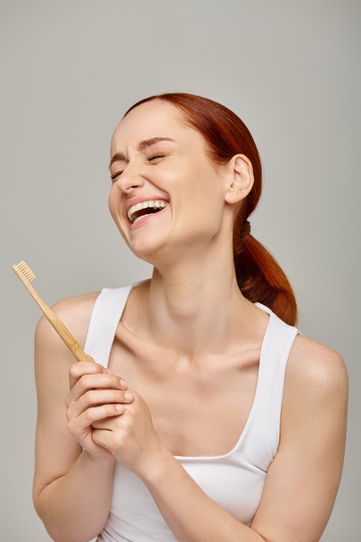 boldog vörös hajú nő tartály tetején kezében fa fogkefe és mosolygós kamera szürke háttér - Fotó, kép
