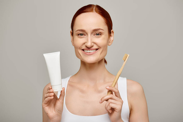 happy redhead woman in tank top holding toothpaste and toothbrush and smiling at camera on grey - Photo, Image