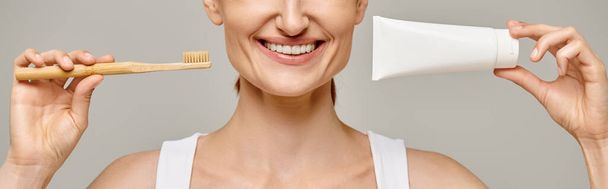 bijgesneden banner van vrolijke vrouw met tandpasta en tandenborstel en glimlachend naar de camera op grijs - Foto, afbeelding