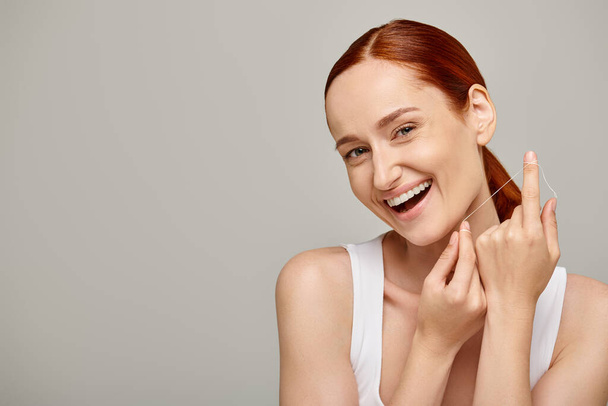 aufgeregtes rothaariges Model mit Zahnseide und Lächeln auf grauem Hintergrund, das für Mundhygiene wirbt - Foto, Bild