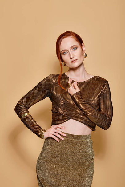 redhead woman in her 30s posing in golden attire and accessories on beige backdrop, hand on hip - Photo, Image