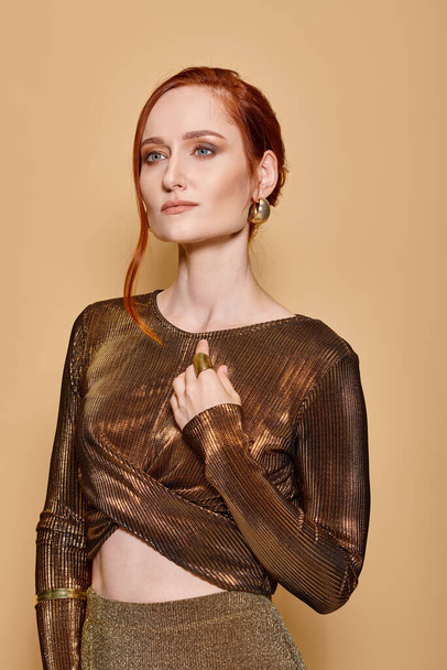 Elegant and redhead woman in her 30s posing in golden attire and accessories on beige backdrop - Фото, зображення