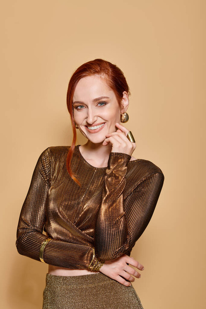 happy and redhead woman in her 30s posing in golden attire and accessories on beige backdrop - 写真・画像