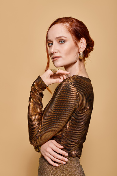 redhead woman in her 30s posing in elegant attire with golden accessories on beige backdrop - Photo, Image