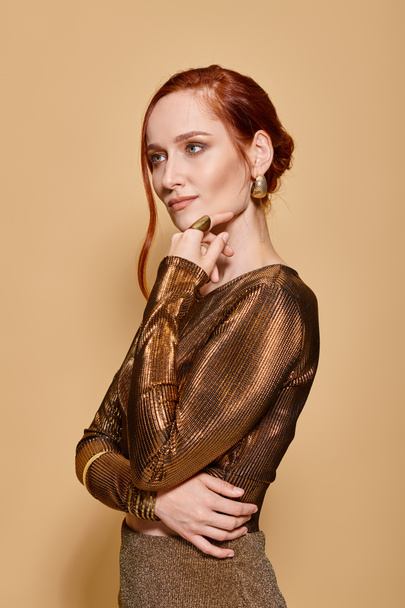 pensive redhead woman in her 30s posing in elegant attire with golden accessories on beige backdrop - Photo, Image