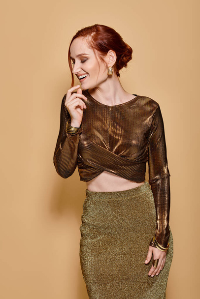 happy redhead woman in her 30s posing in elegant attire and golden accessories on beige backdrop - Photo, Image