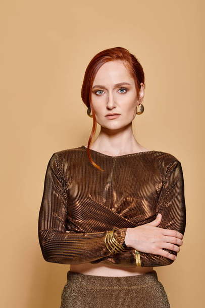 redhead woman in her 30s posing in trendy outfit with golden accessories on beige backdrop, portrait - Photo, Image