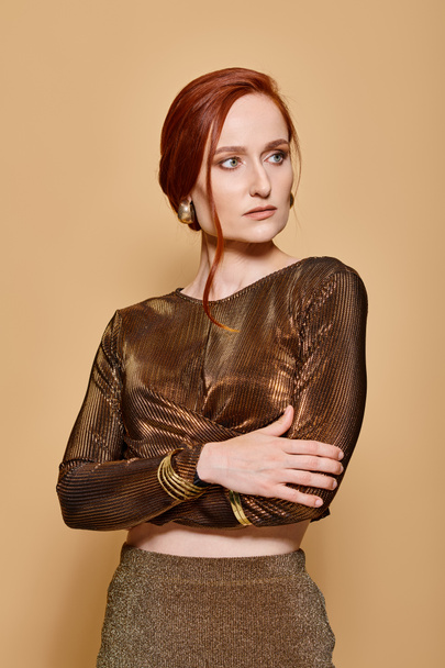 redhead woman in her 30s posing in stylish outfit with golden accessories on beige background - Photo, Image