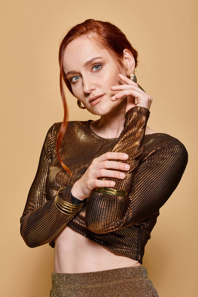 redhead woman in her 30s posing in stylish outfit and golden accessories with hand near face - Photo, Image