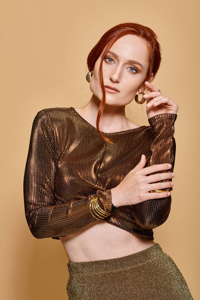 redhead woman in her 30s posing in stylish attire and golden accessories with hands near face - Photo, Image