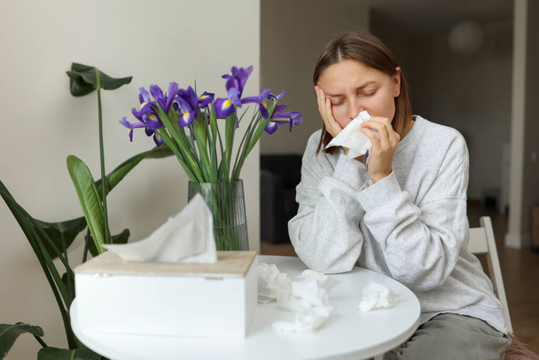 Nemocná nezdravá alergická mladá žena zakrývající nos papírovou tkání má ucpaný nos, kýchá z pylu květin duhovky v kuchyni doma. Dívka s chřipkou, svěděním nebo kašlem, sezónní alergií, rýmou - Fotografie, Obrázek