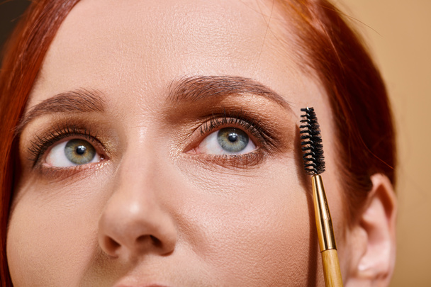 cropped photo of redhead woman with green eyes holding eye brow brush on beige backdrop - Photo, Image