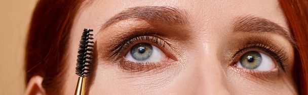 close up photo of redhead woman with green eyes applying mascara on beige background, banner - Photo, Image