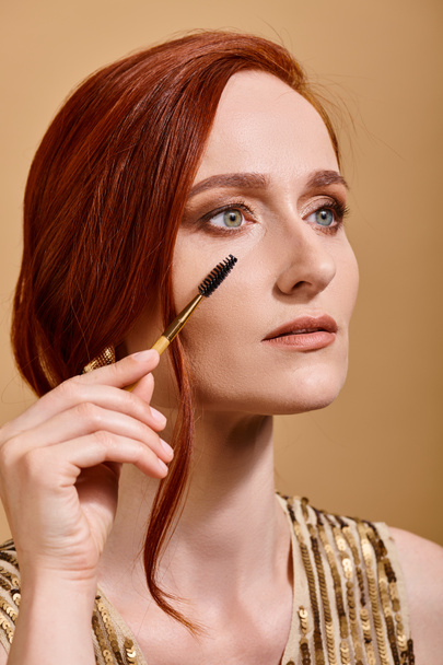 portret van roodharige vrouw met groene ogen aanbrengen mascara op beige achtergrond, make-up schoonheid - Foto, afbeelding