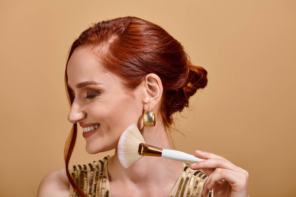 mujer pelirroja feliz en vestido de lentejuelas de oro con cepillo de maquillaje sobre fondo beige, mirada de belleza - Foto, imagen