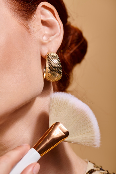 mujer pelirroja recortada en pendiente de oro aplicando bronceador con cepillo de maquillaje sobre fondo beige - Foto, imagen