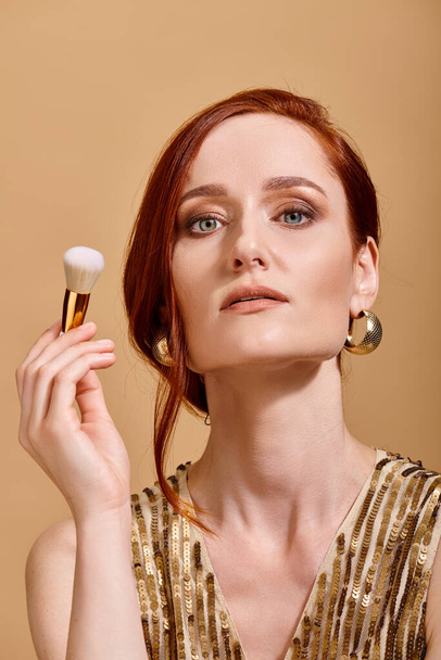 zelfverzekerde roodharige vrouw in goud oorbellen houden make-up borstel op beige achtergrond, schoonheid concept - Foto, afbeelding