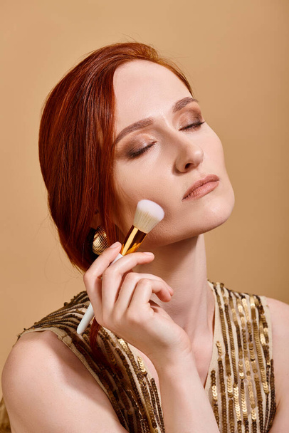 mujer pelirroja en pendiente de oro aplicando la base de la cara con el cepillo de maquillaje sobre fondo beige - Foto, imagen