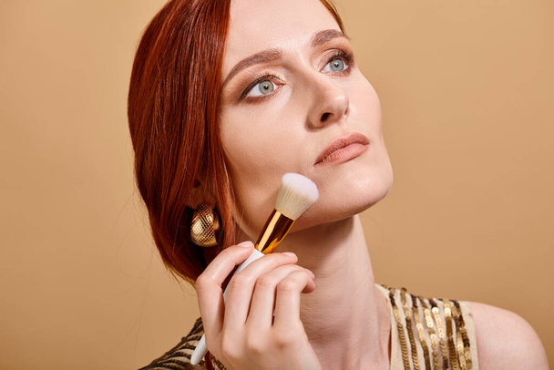 rousse femme en boucle d'oreille en or appliquant fond de teint visage avec pinceau de maquillage sur fond beige - Photo, image