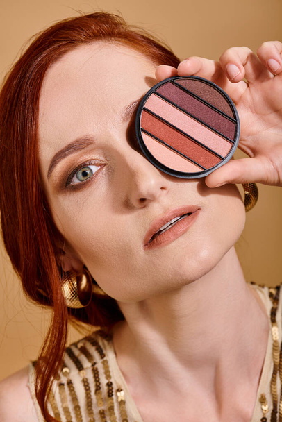 roodharige vrouw bedekt haar gezicht met oogschaduw palet op beige achtergrond, schoonheid concept - Foto, afbeelding