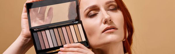 mujer pelirroja sosteniendo la paleta de sombra de ojos cerca de la cara sobre fondo beige, banner de publicidad de maquillaje - Foto, imagen