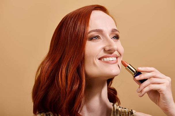 Mulher alegre e ruiva sorrindo e aplicando batom nu no fundo bege, produto de maquiagem - Foto, Imagem