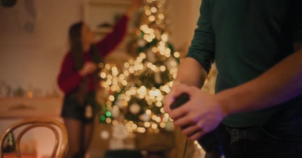Man connecting plug of led light on Christmas tree by female friend in living room - Footage, Video