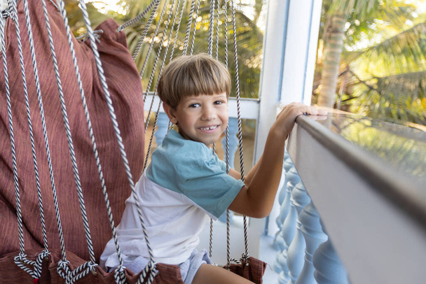 Portret van een gelukkige jongen tijdens de zomervakantie, op zee in het hotel, die zit in een schommelstoel op een open balkon en glimlacht, kijkend naar de camera. Recreatie en reizen. Verzekering en kinderbescherming. - Foto, afbeelding