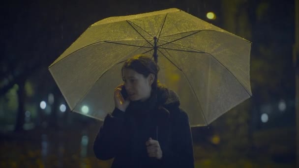 Mujer triste hablando en el teléfono celular fuera por la noche en tiempo de lluvia - Imágenes, Vídeo