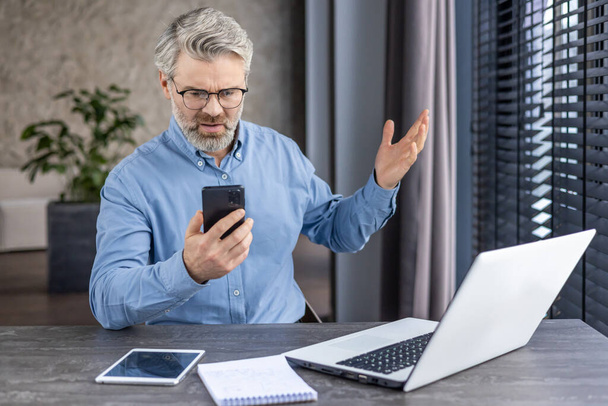 İş yerindeki patron üzgün iş adamına telefonda online mesaj geldi. Yetişkin, yaşlı, gri saçlı bir adam akıllı telefonunda uygulama kullanıyor.. - Fotoğraf, Görsel