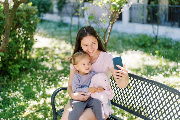 彼女の腕に小さな女の子と笑顔の母親は,庭のベンチに座っている間,スマートフォンでセルフィーを取ります. 高品質の写真 - 写真・画像