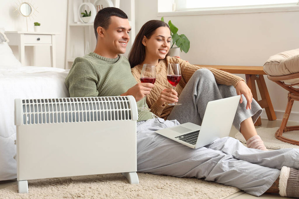 Молодая пара с вином и ноутбуком согреваются возле радиатора в спальне - Фото, изображение