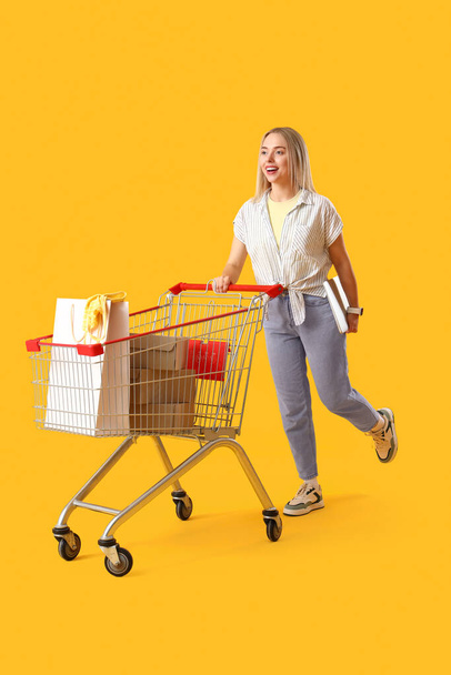 黄色い背景のショッピングバッグでいっぱいのラップトップとマーケットトロリーを持つ若い女性 - 写真・画像
