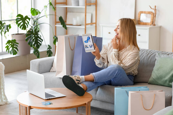 Młoda kobieta z laptopem, tabletem i torbami na zakupy siedzi na kanapie w domu - Zdjęcie, obraz