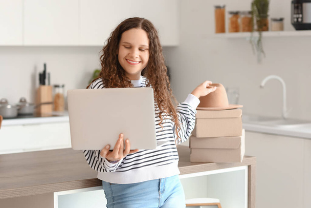 Νεαρή γυναίκα με φορητό υπολογιστή και κουτιά ψώνια σε απευθείας σύνδεση στην κουζίνα - Φωτογραφία, εικόνα