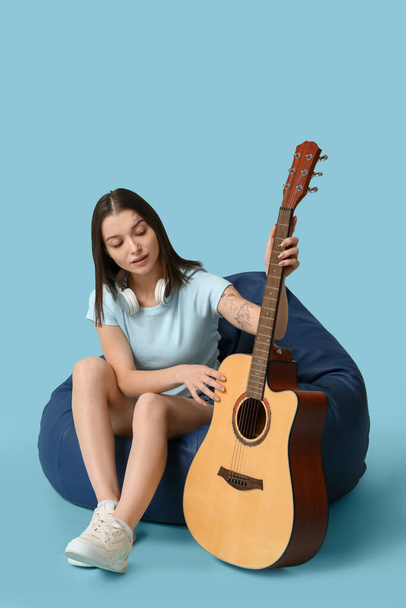 Όμορφη νεαρή γυναίκα με ακουστική κιθάρα κάθεται σε pouf σε μπλε φόντο - Φωτογραφία, εικόνα