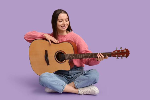 ライラックの背景に座っているアコースティックギターを持つかなり若い女性 - 写真・画像