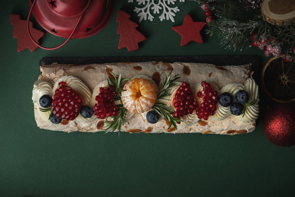 Baiser-Rolle, bestreut mit Puderzucker, dekoriert mit Blaubeeren, Granatapfel und Mandelblättchen auf dem dunkelgrünen weihnachtlichen Hintergrund. Flache Lage - Foto, Bild